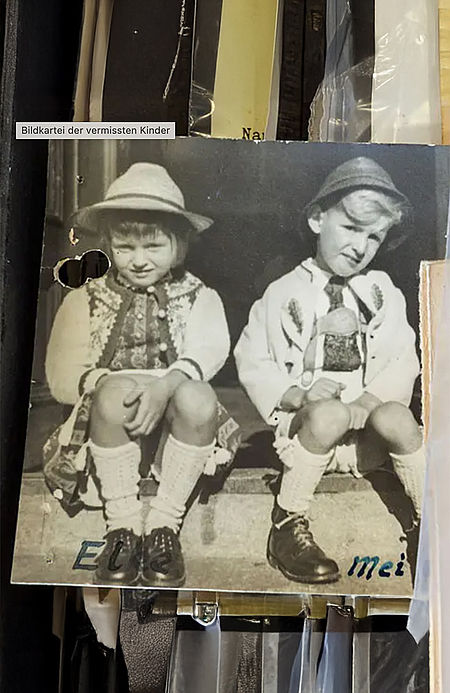 Zeitzeugen_01 Schwarz-weiß-Foto aus der Bildkartei vermisster Kinder: Zwei kleine Kinder in traditioneller Tracht sitzen nebeneinander auf einer Treppe. Die Karte zeigt Gebrauchsspuren und ist handbeschriftet mit den Namen „Els“ und „Mei“.